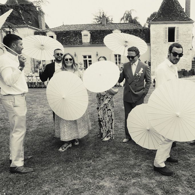 Wedding guests holding white paper parasols on a lawn in front of a French château