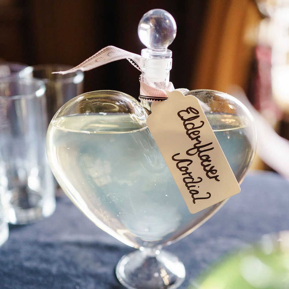 Heart-shaped glass decanter of elderflower cordial with a hand-lettered tag and ribbon