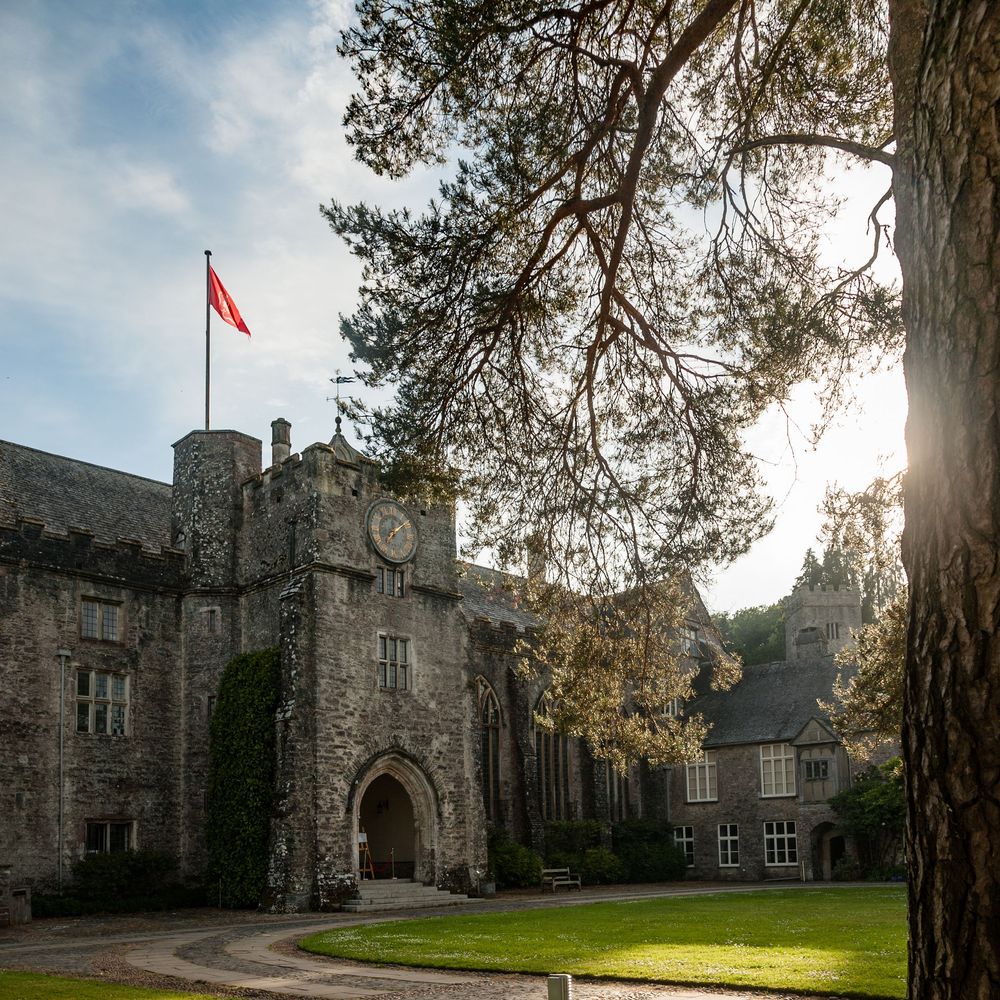 Historic stone manor with clock tower and red flag overlooking a sunlit lawn and gardens