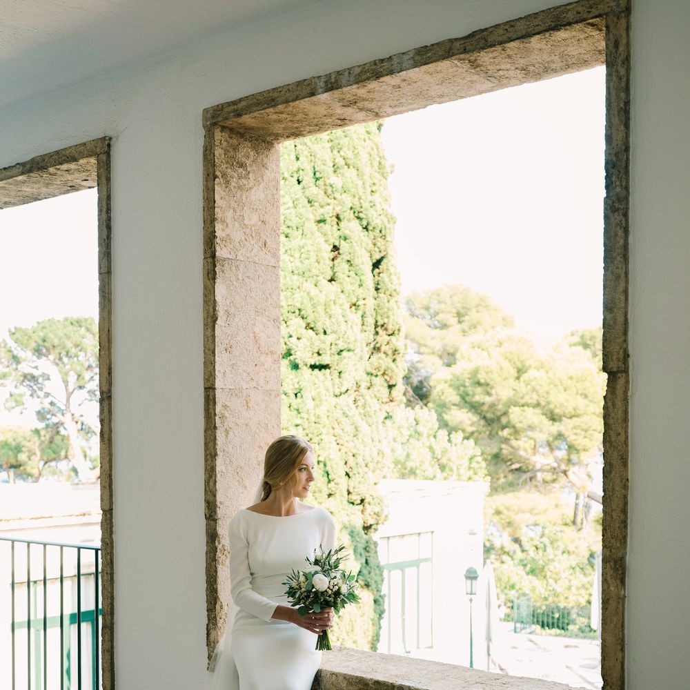 Bride in a long-sleeved satin gown holding a bouquet, seated by a large stone window
