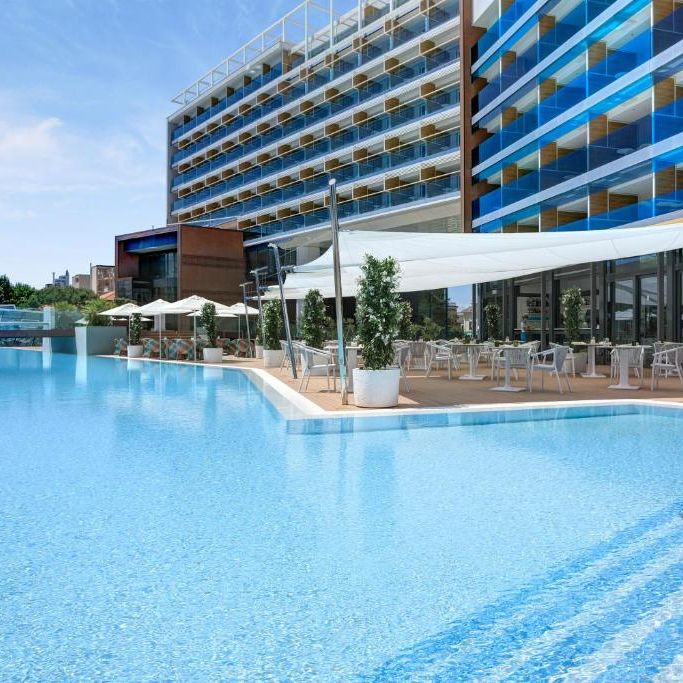 Outdoor pool with white parasols, dining terrace and potted olive trees beside a modern hotel
