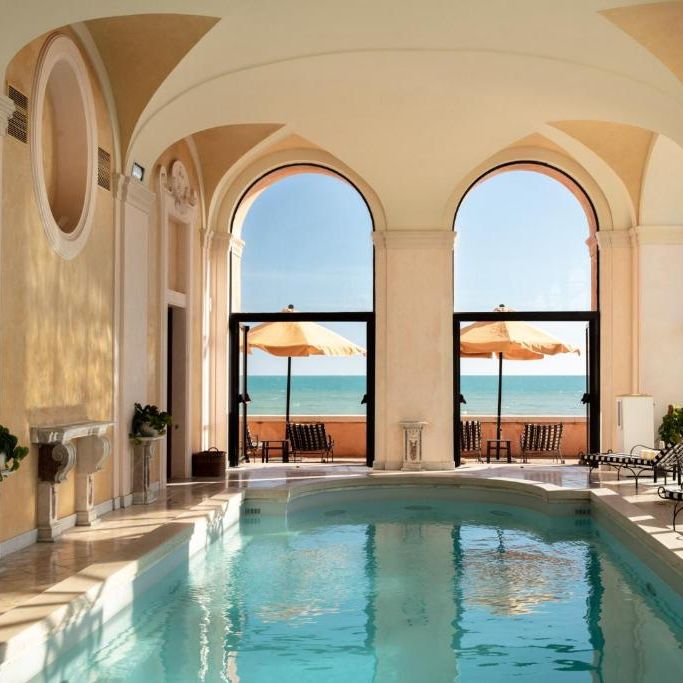 Indoor pool beneath vaulted ceilings with arched windows framing a sea view and parasols on the terrace