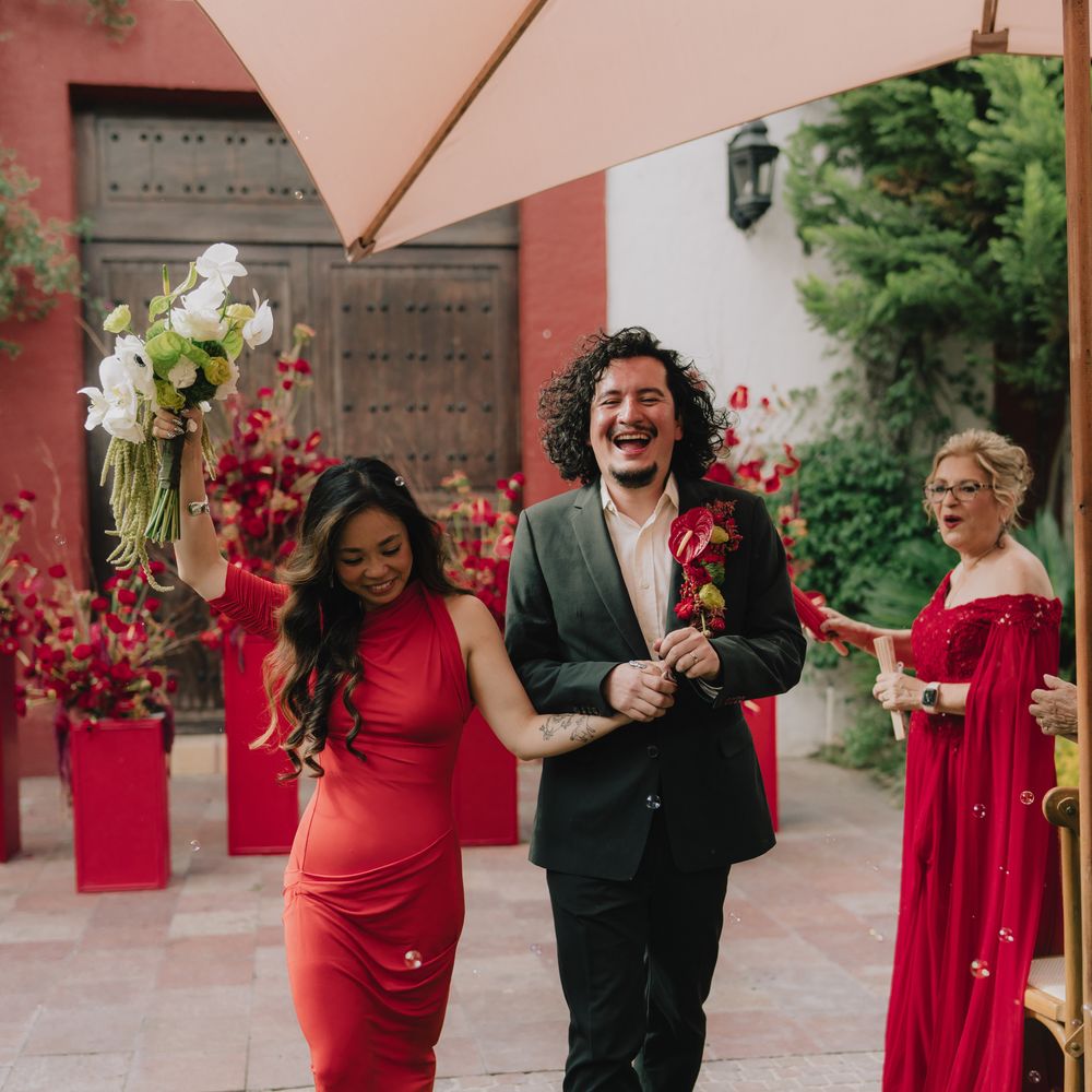 Couple in red and green celebrate walking through a courtyard beneath a blush umbrella