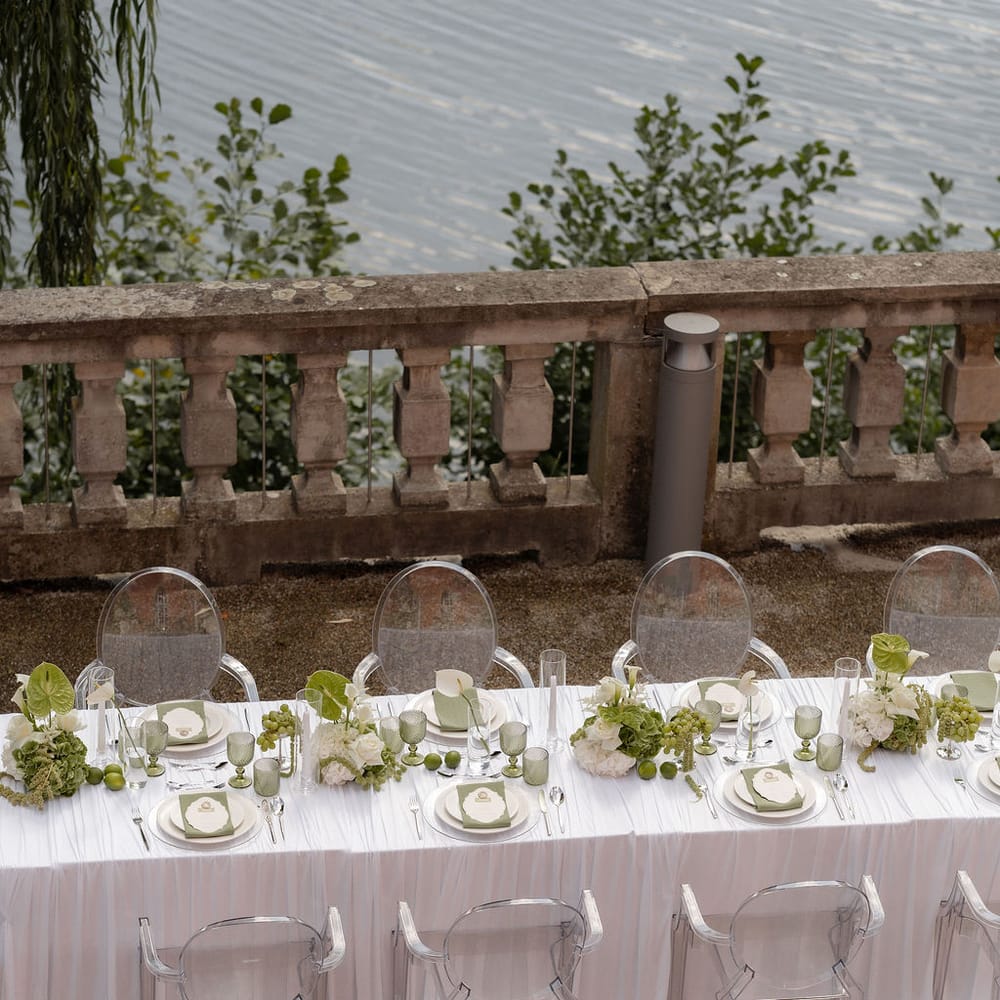 Lakeside wedding table with ghost chairs, green glassware and white floral styling by a stone balustrade
