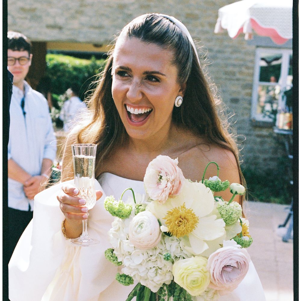 Laughing bride in off-shoulder gown holds champagne and a pastel ranunculus bouquet outdoors