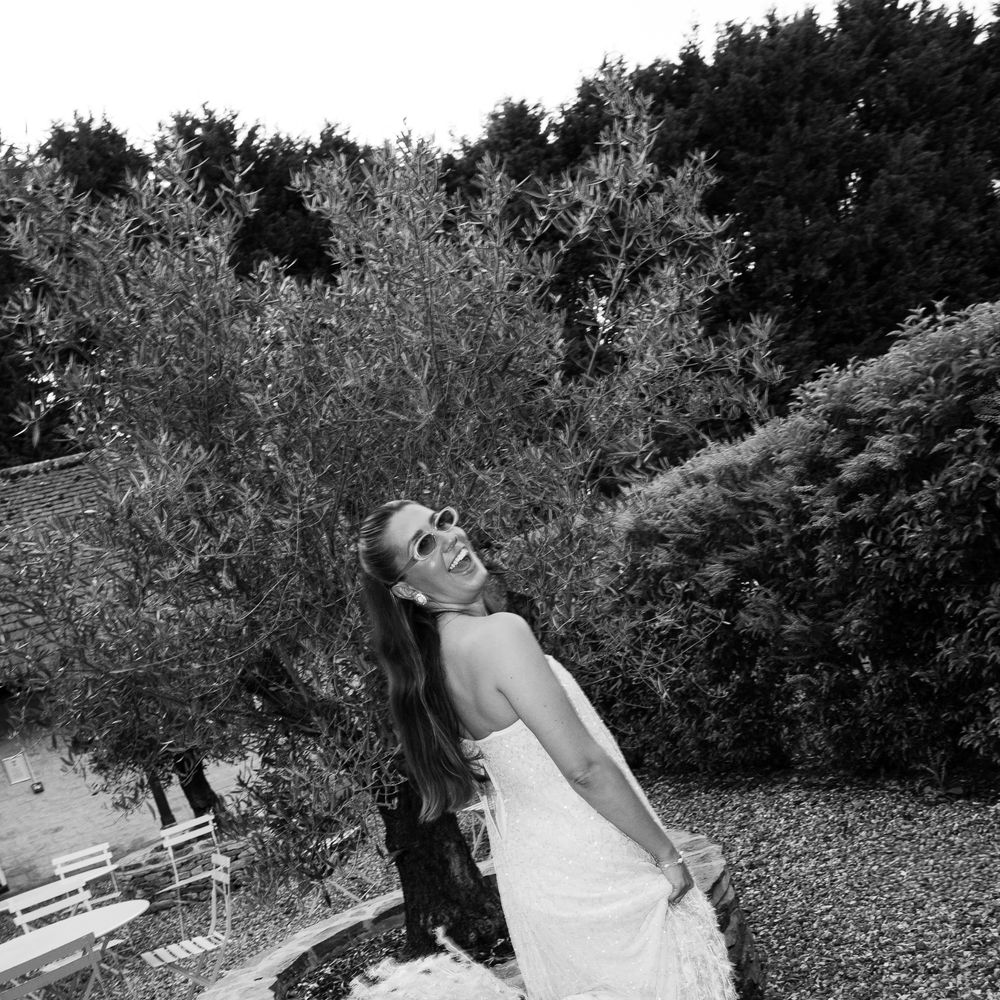 Laughing bride in fringed sequin gown and sunglasses beside a stone well in a garden