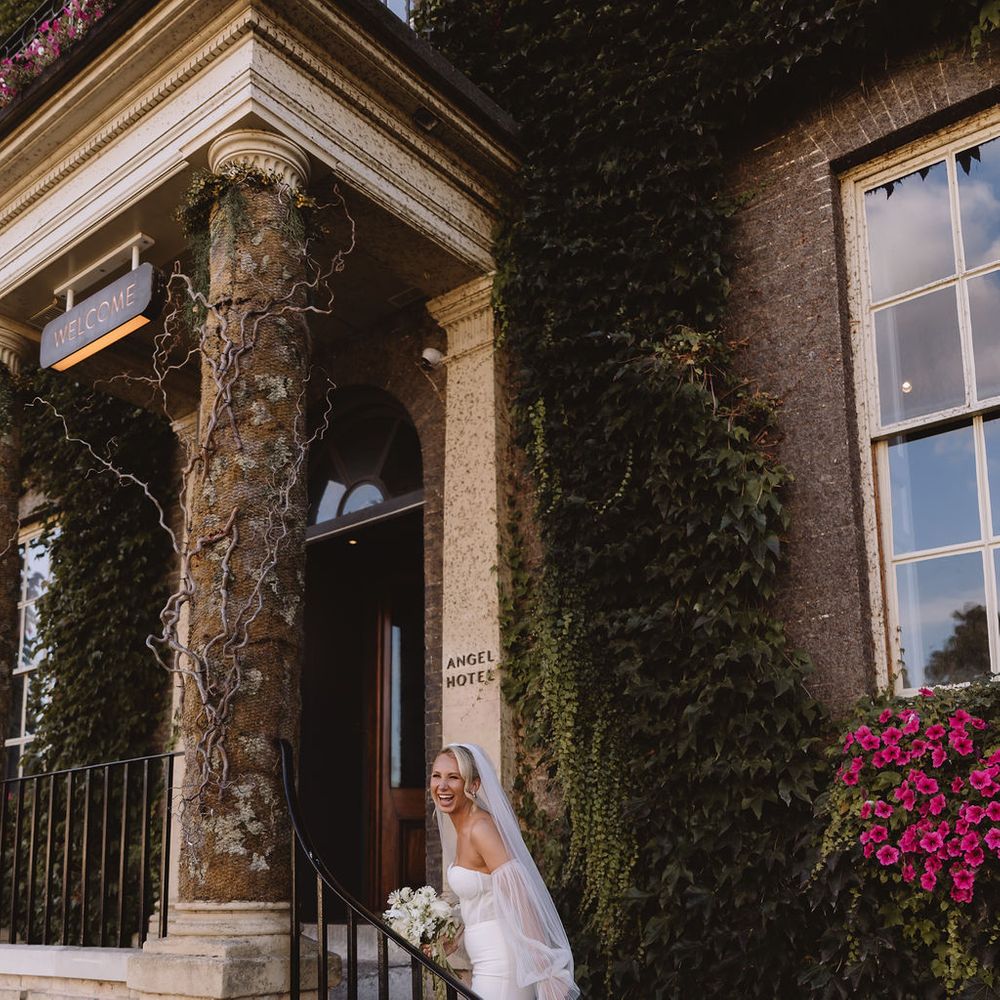 Laughing bride in a strapless gown descends ivy-clad hotel steps holding a white bouquet.