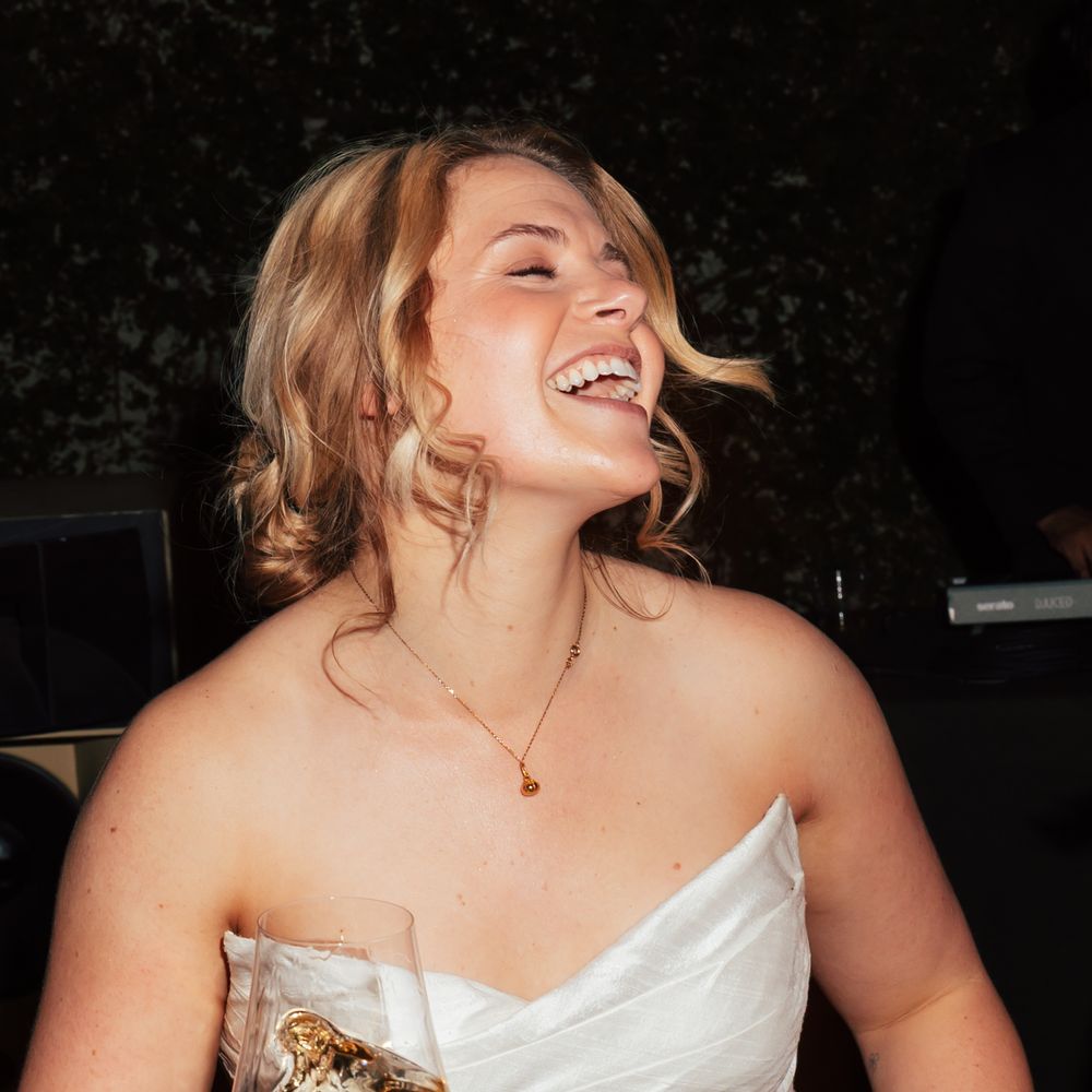 Laughing bride in a strapless satin gown holding a glass of white wine at her evening reception
