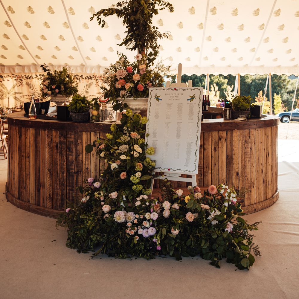 Rustic wooden bar in a marquee framed by a pole draped with abundant seasonal florals and a table plan