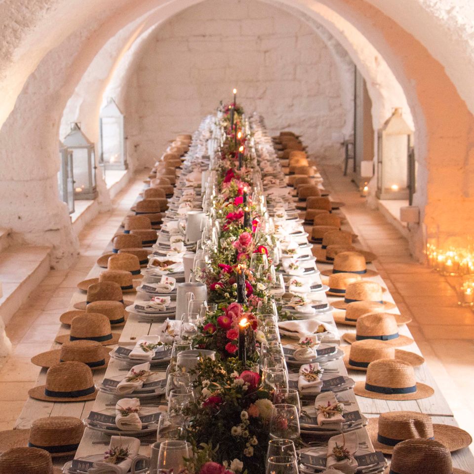 Long banquet table with straw hat favours and pink floral runner in a whitewashed vaulted room