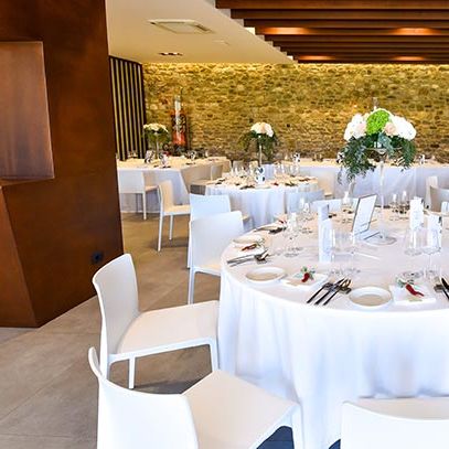 Round tables dressed in white linen with tall floral centrepieces in a stone-walled dining room