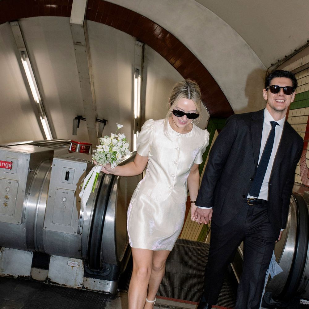 Newlywed couple in sunglasses hold hands stepping off an escalator in a London Underground station