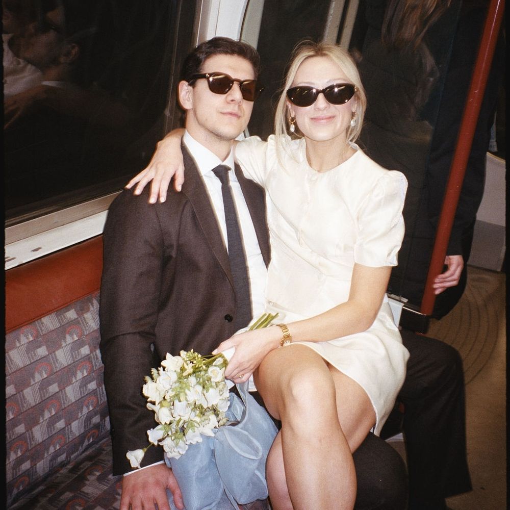 Newlywed couple in sunglasses sit close together on a London Underground train with a white bouquet