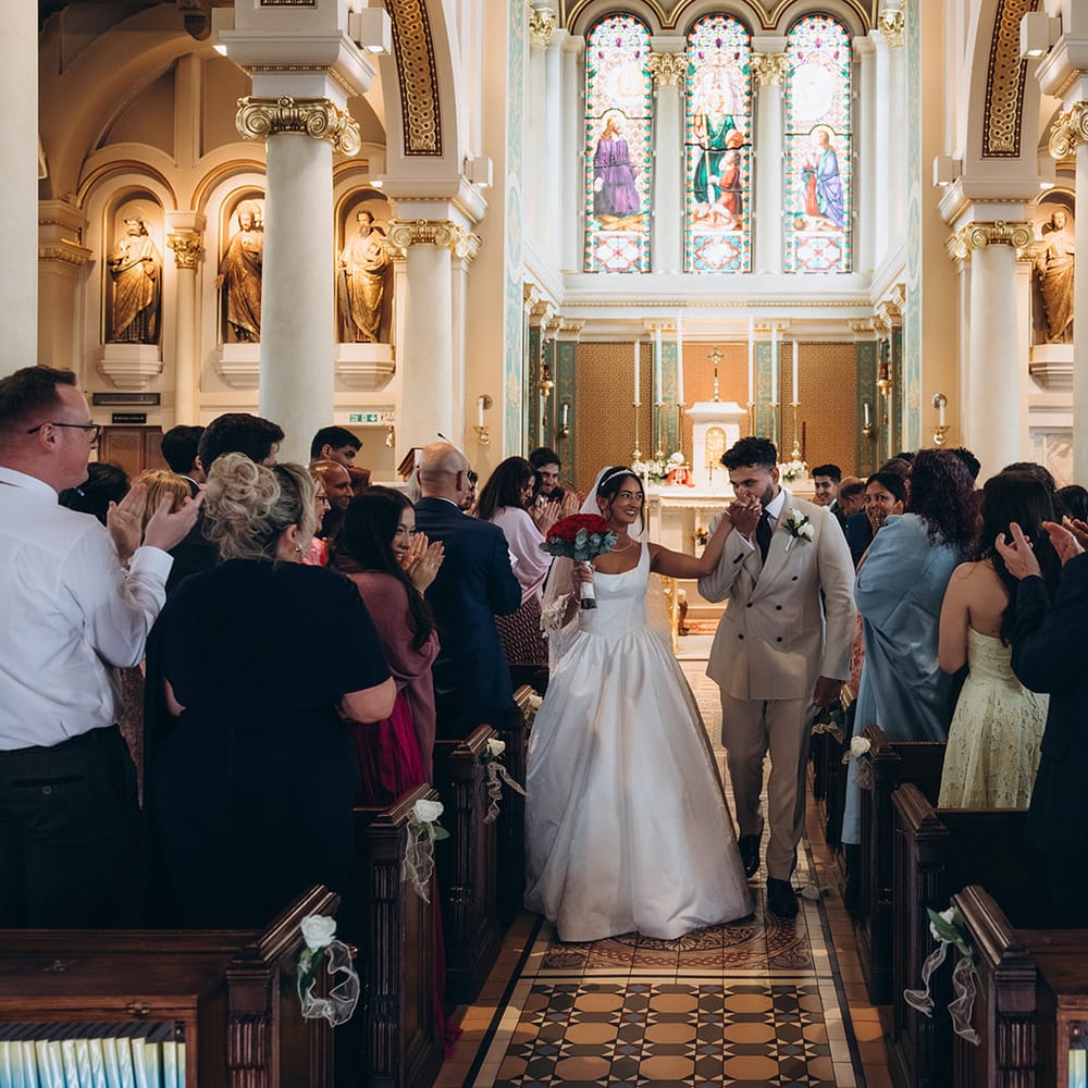 Newlyweds walk down the aisle as guests applaud beneath stained glass windows in an ornate church