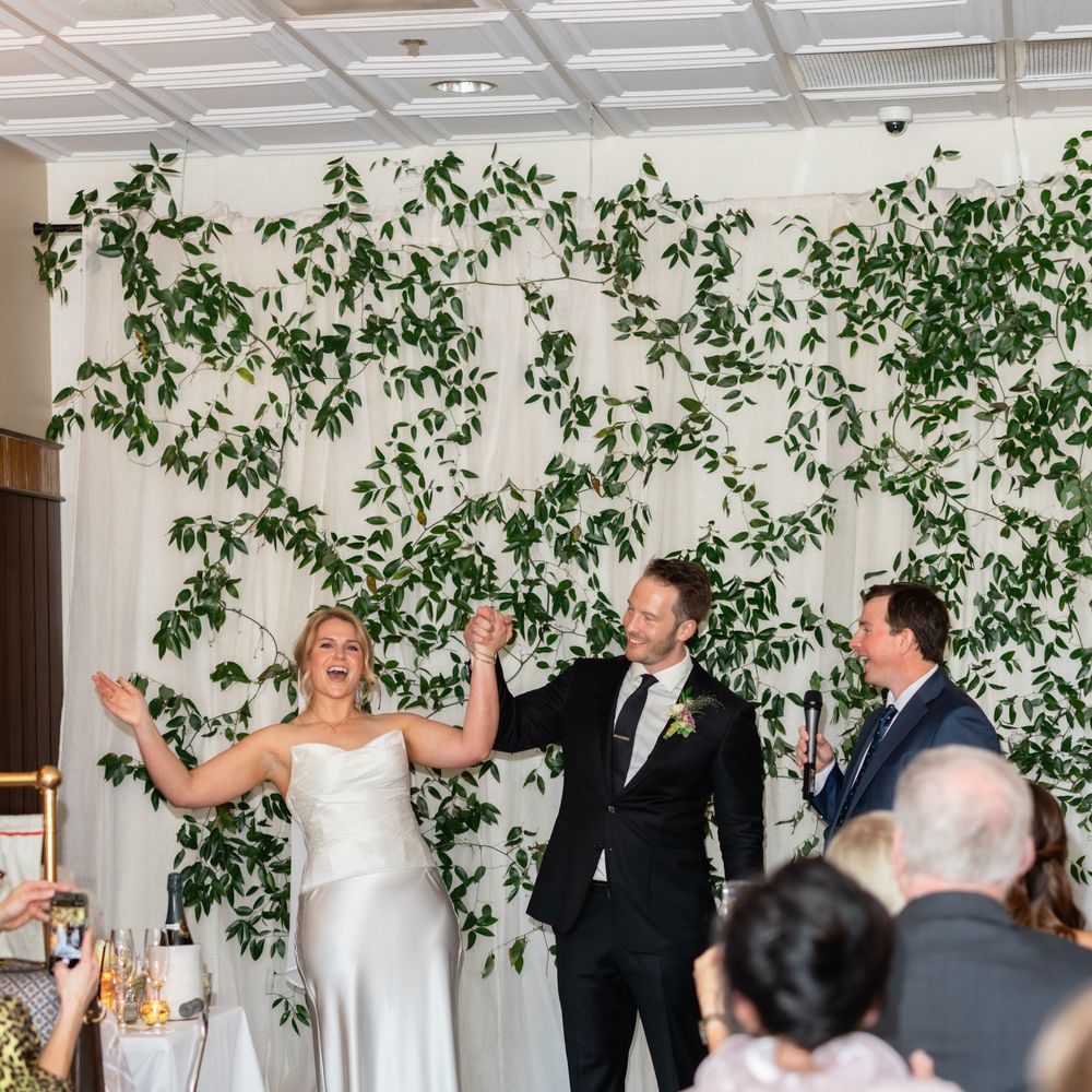 Bride and groom raise clasped hands in celebration before a foliage-draped backdrop as guests look on