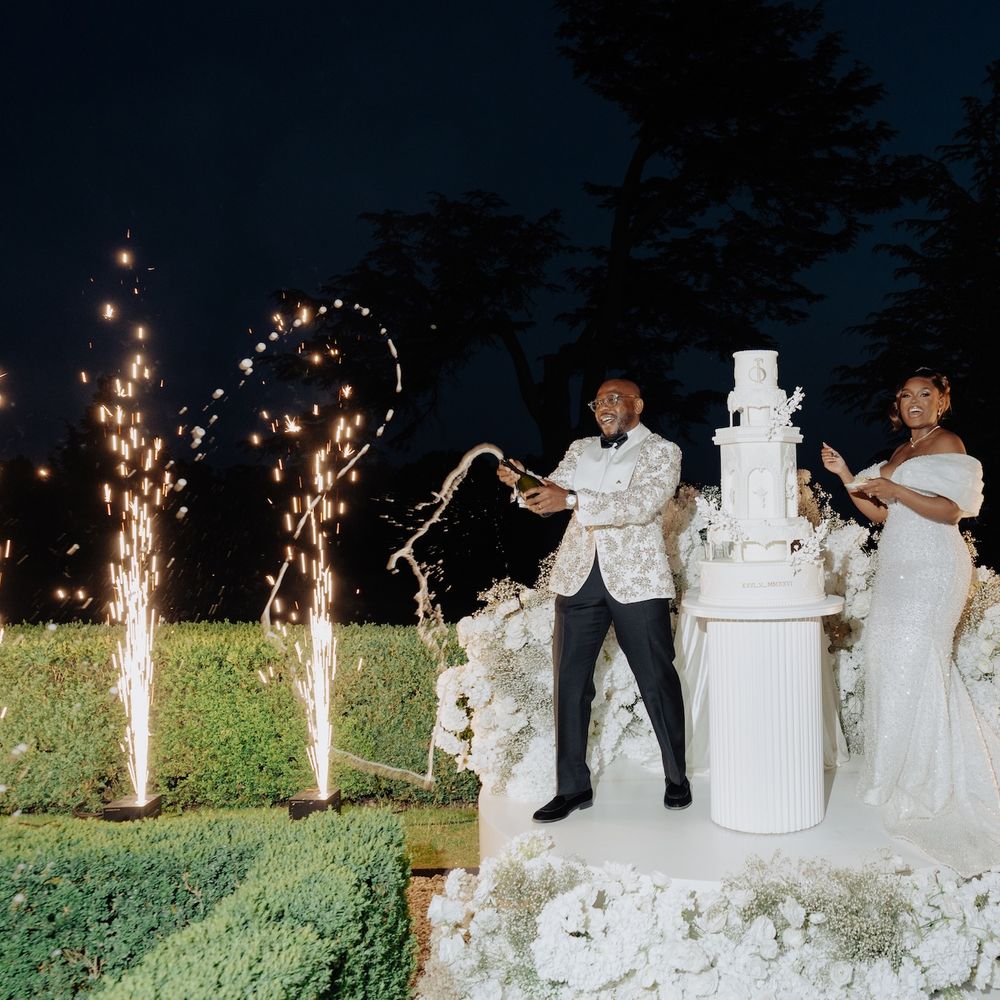 Bride and groom celebrate beside a tall white wedding cake with sparkler fountains and champagne at night