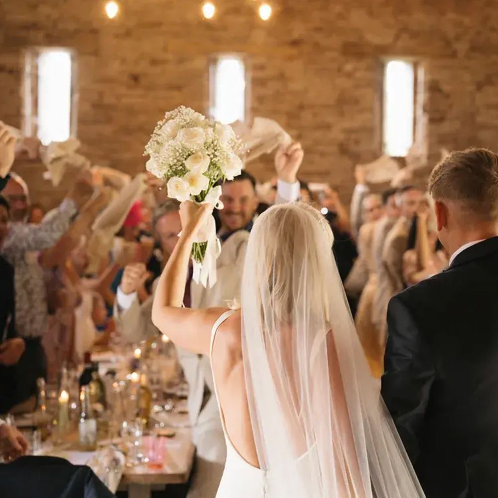 Bride and groom raise arms in celebration as guests wave napkins in a rustic stone barn