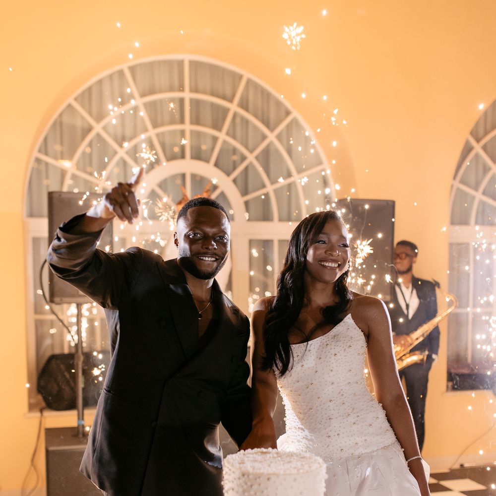 Newlyweds smiling beside a two-tier pearl-detailed cake with sparklers and a saxophonist behind