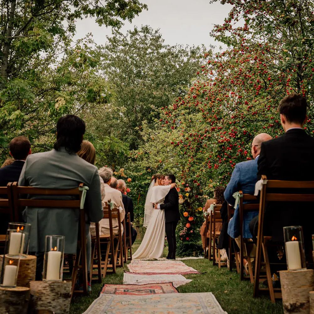 Couple share first kiss at outdoor orchard ceremony lined with candles and vintage rugs