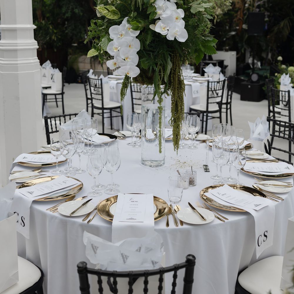 Round wedding table with tall white orchid centrepiece, gold charger plates and glassware in a glasshouse