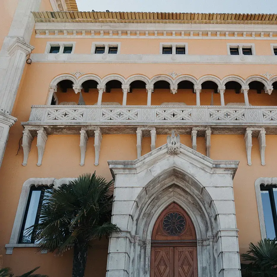 Ornate terracotta villa facade with carved stone balcony, arched doorway and palm trees