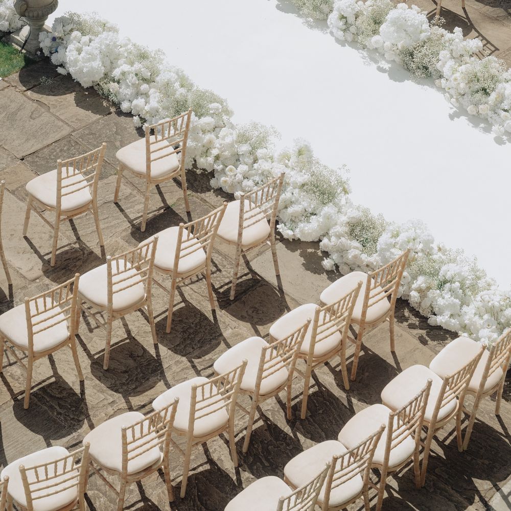 White aisle runner lined with white blooms and stone urns between rows of Chiavari chairs outdoors