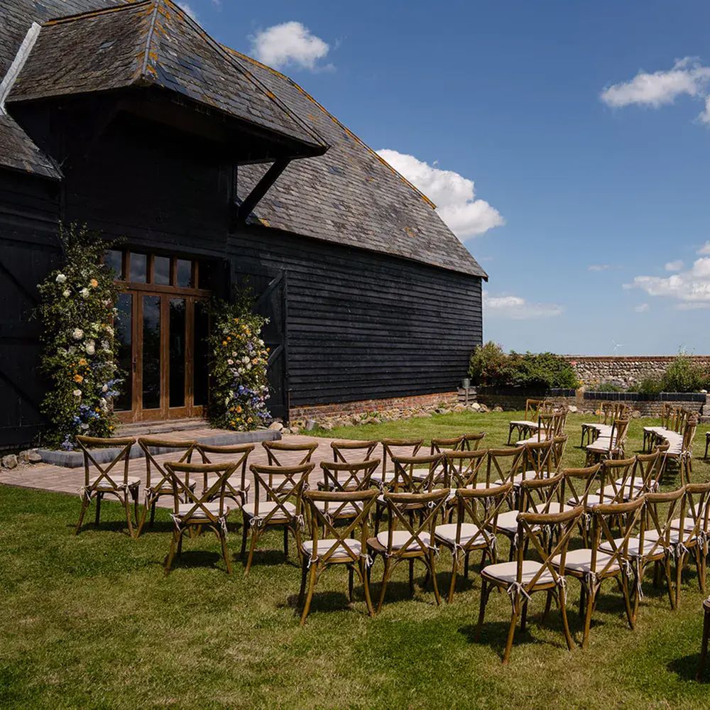 Wooden cross-back chairs arranged in curves for an outdoor barn ceremony beneath blue skies