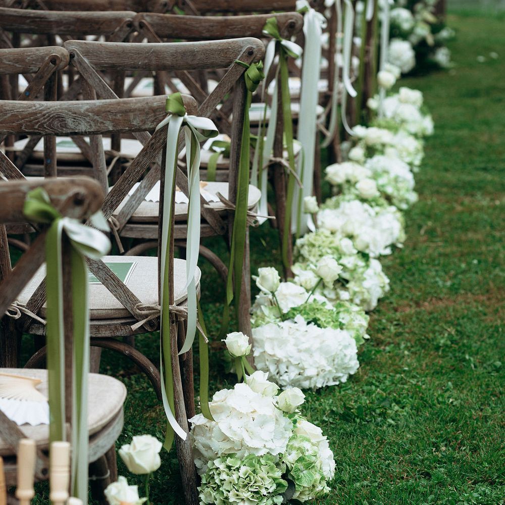Rustic wooden chairs with green ribbons beside white hydrangeas lining a grass ceremony aisle