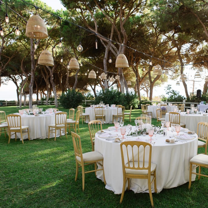 Outdoor wedding reception with round tables, gold chairs and rattan pendant lights beneath pine trees
