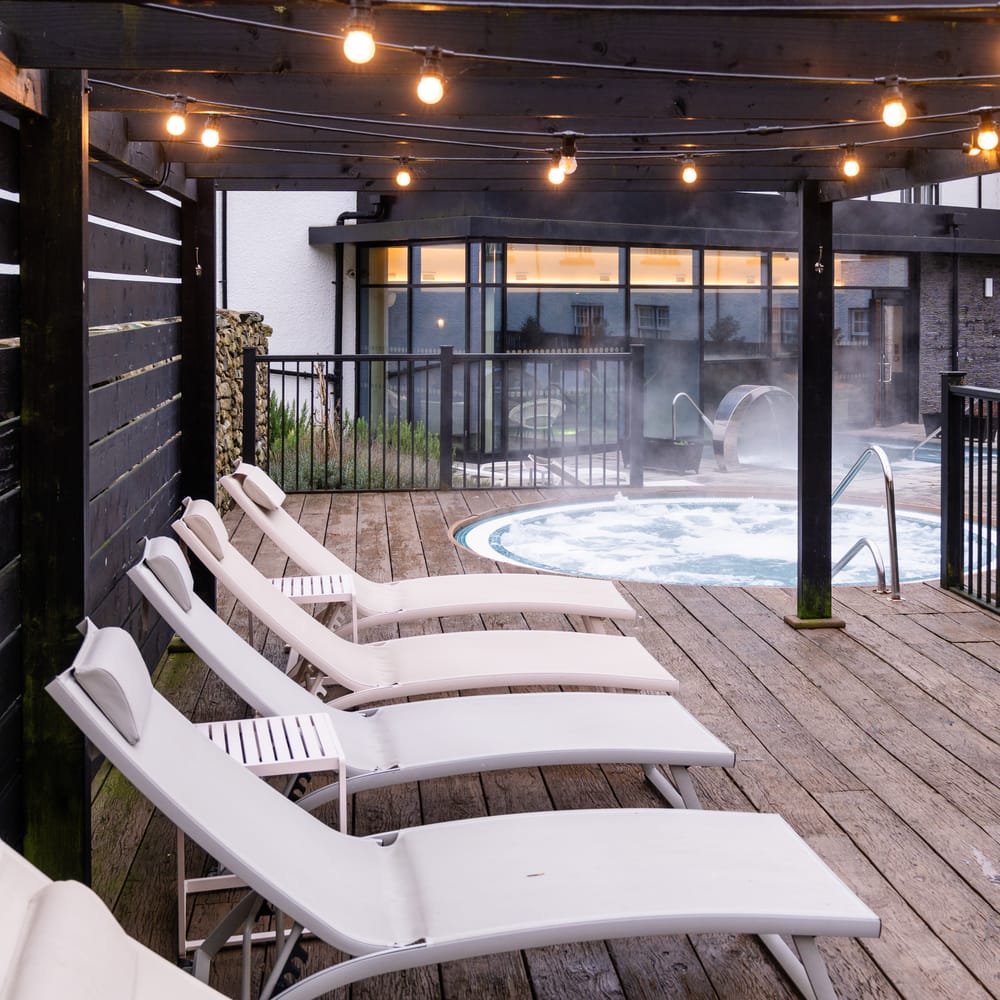 White loungers beside a steaming outdoor hot tub on a decked spa terrace at dusk