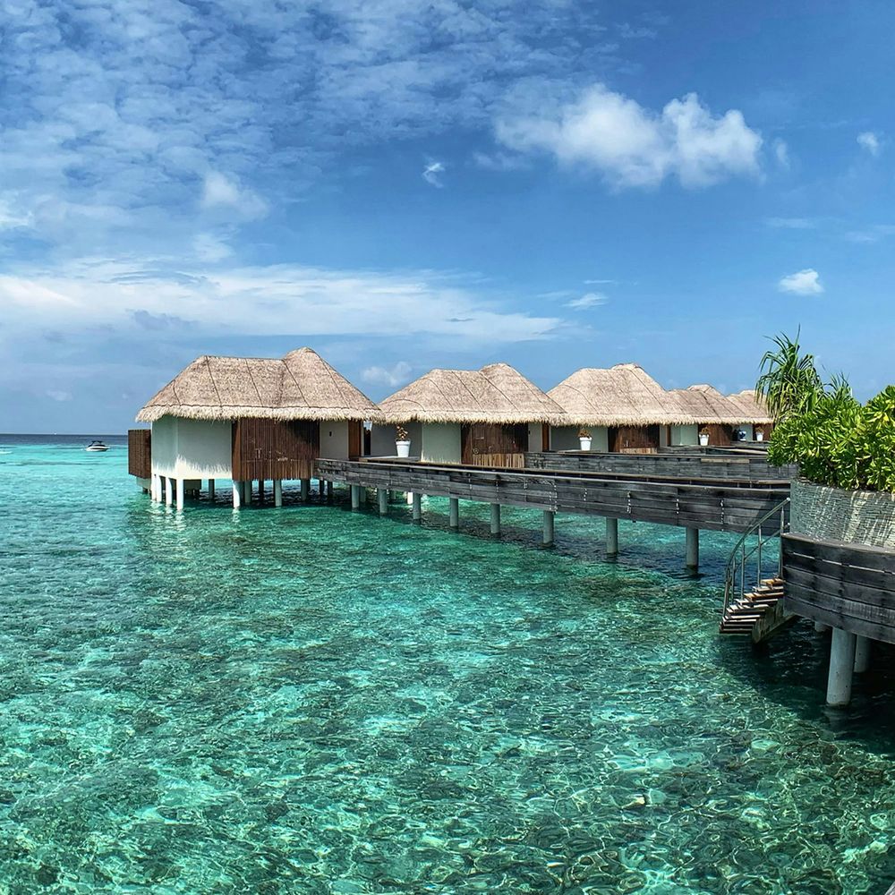 Thatched overwater villas connected by a wooden walkway above clear turquoise lagoon waters.