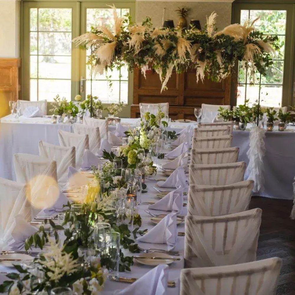 Long banquet tables dressed in white with foliage runners beneath a pampas grass hanging installation