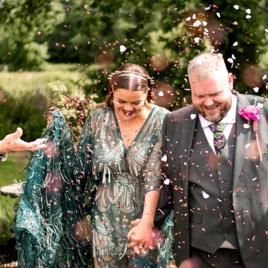 Newlyweds hold hands and smile as guests throw petal confetti in a leafy garden
