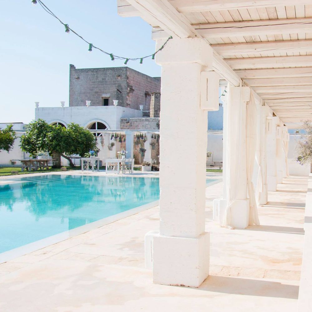 Sunlit poolside terrace with white pillars, drapes and stone masseria buildings beyond
