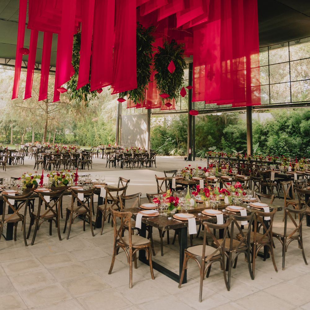 Outdoor reception with suspended red banners, hanging greenery and rustic wooden tables set with red blooms