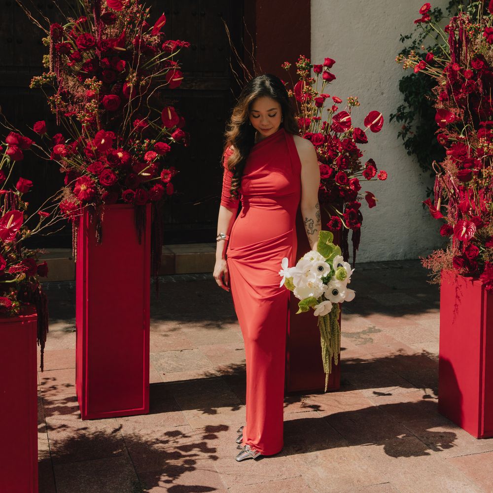 Woman in a red one-shoulder gown holding white anemones beside crimson floral pedestals