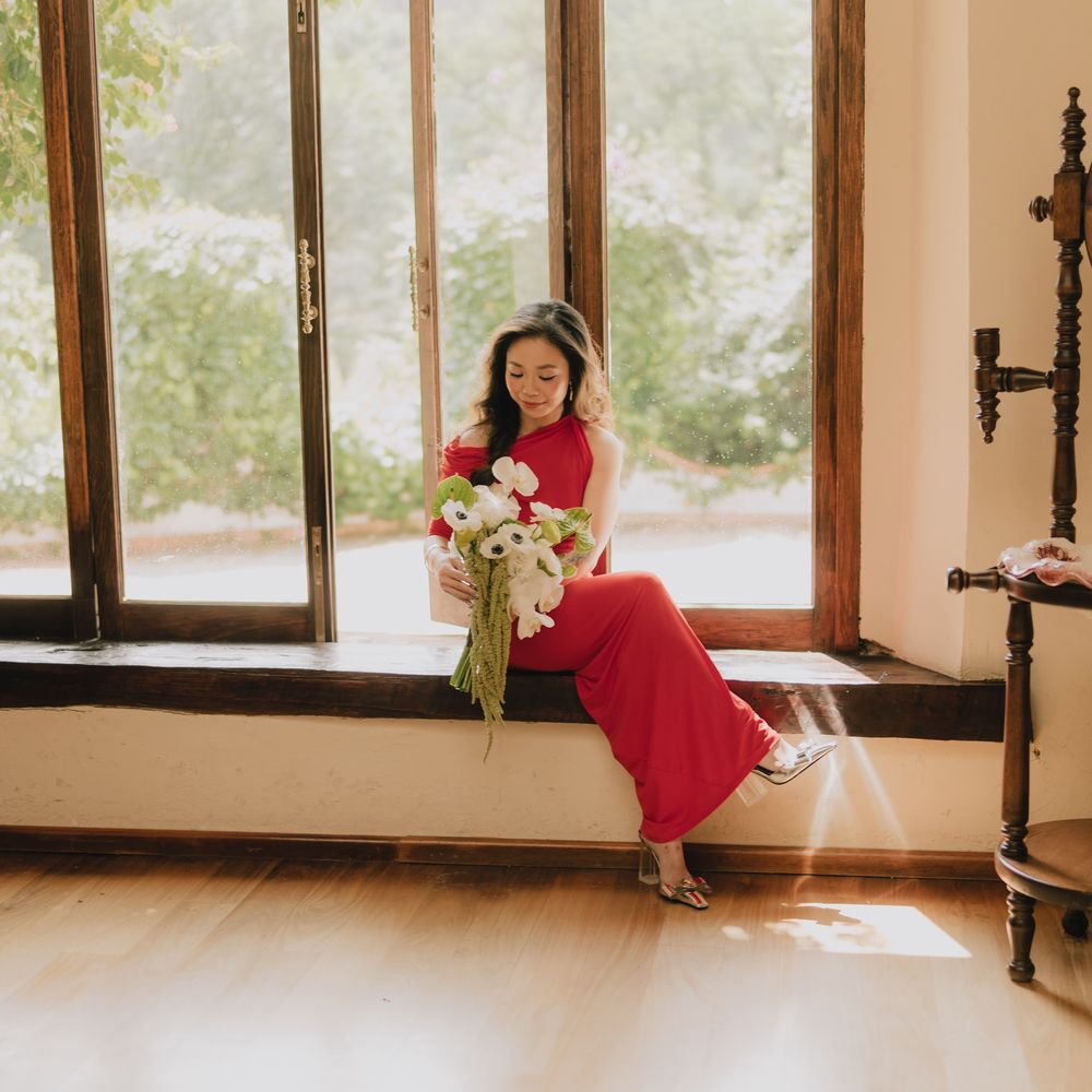 Woman in a red gown sits on a sunlit window seat holding a white anemone bouquet