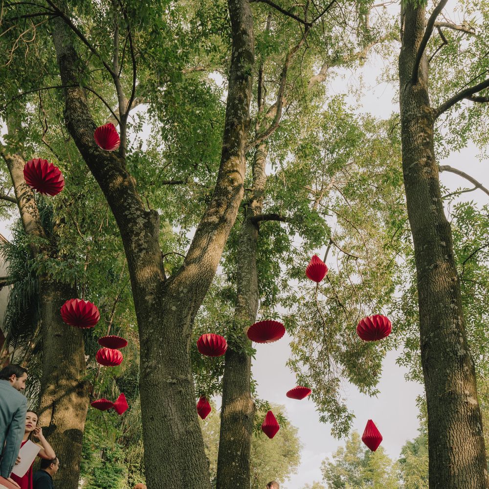 Red paper lanterns hanging from tall trees above guests at an outdoor garden wedding