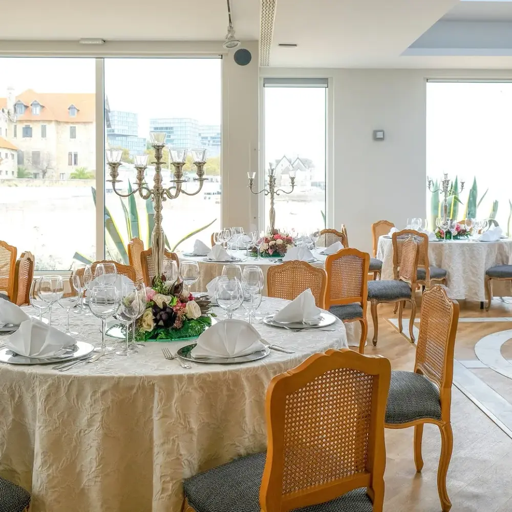 Round reception tables with silver candelabra, floral centrepieces and cane-backed chairs by large windows