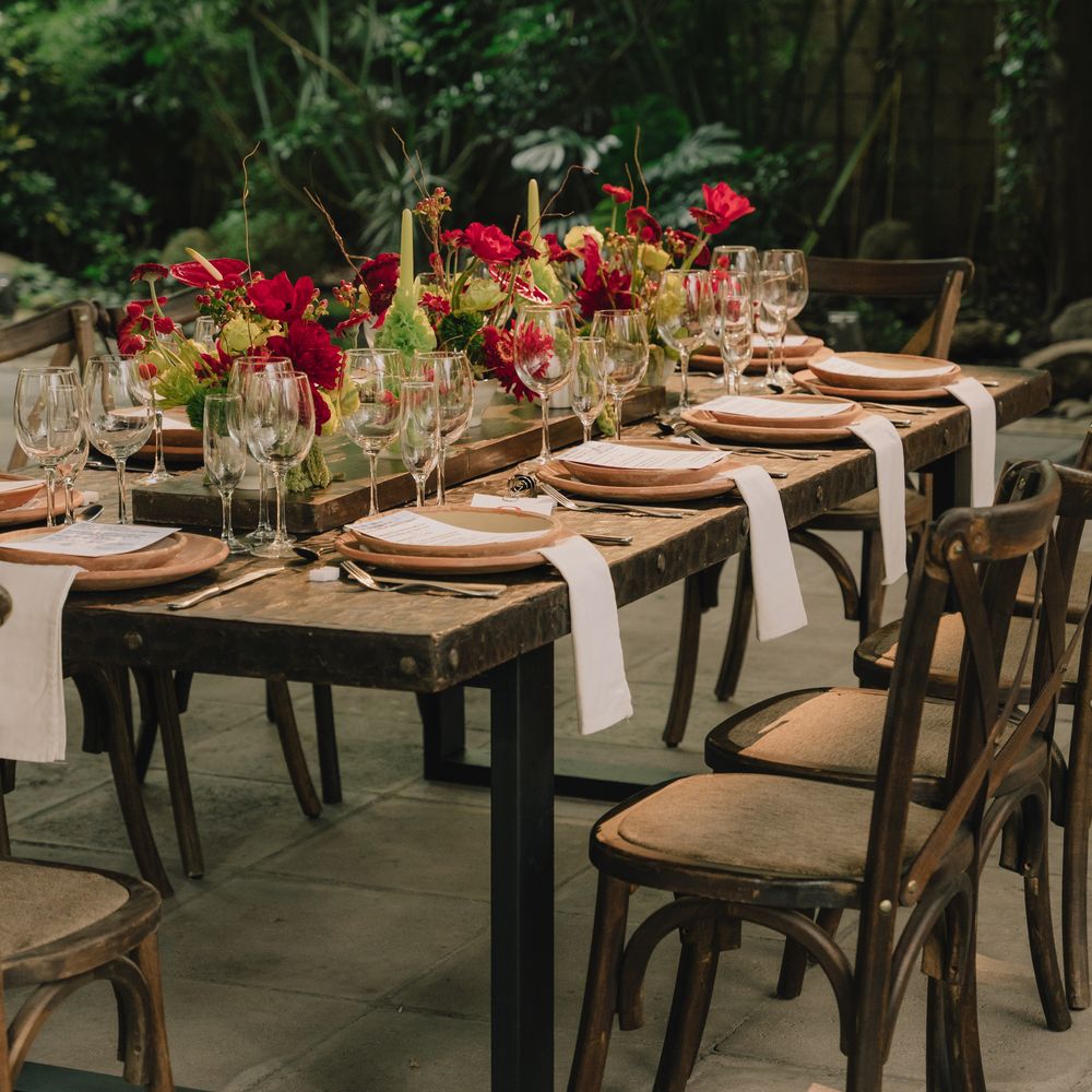 Rustic wooden banquet table set with red florals, wooden chargers and cross-back chairs outdoors