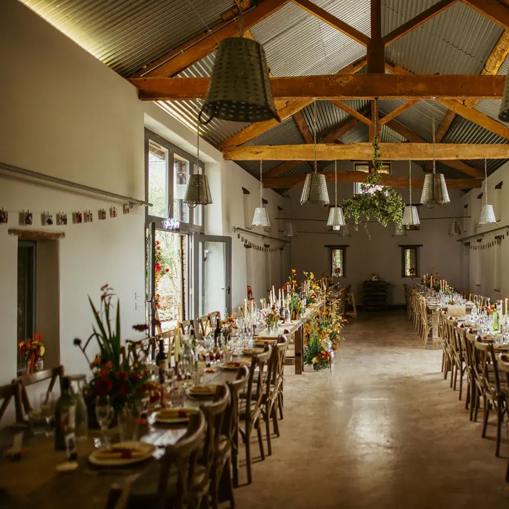 Rustic barn reception with long wooden tables, autumnal flowers and taper candles beneath exposed beams