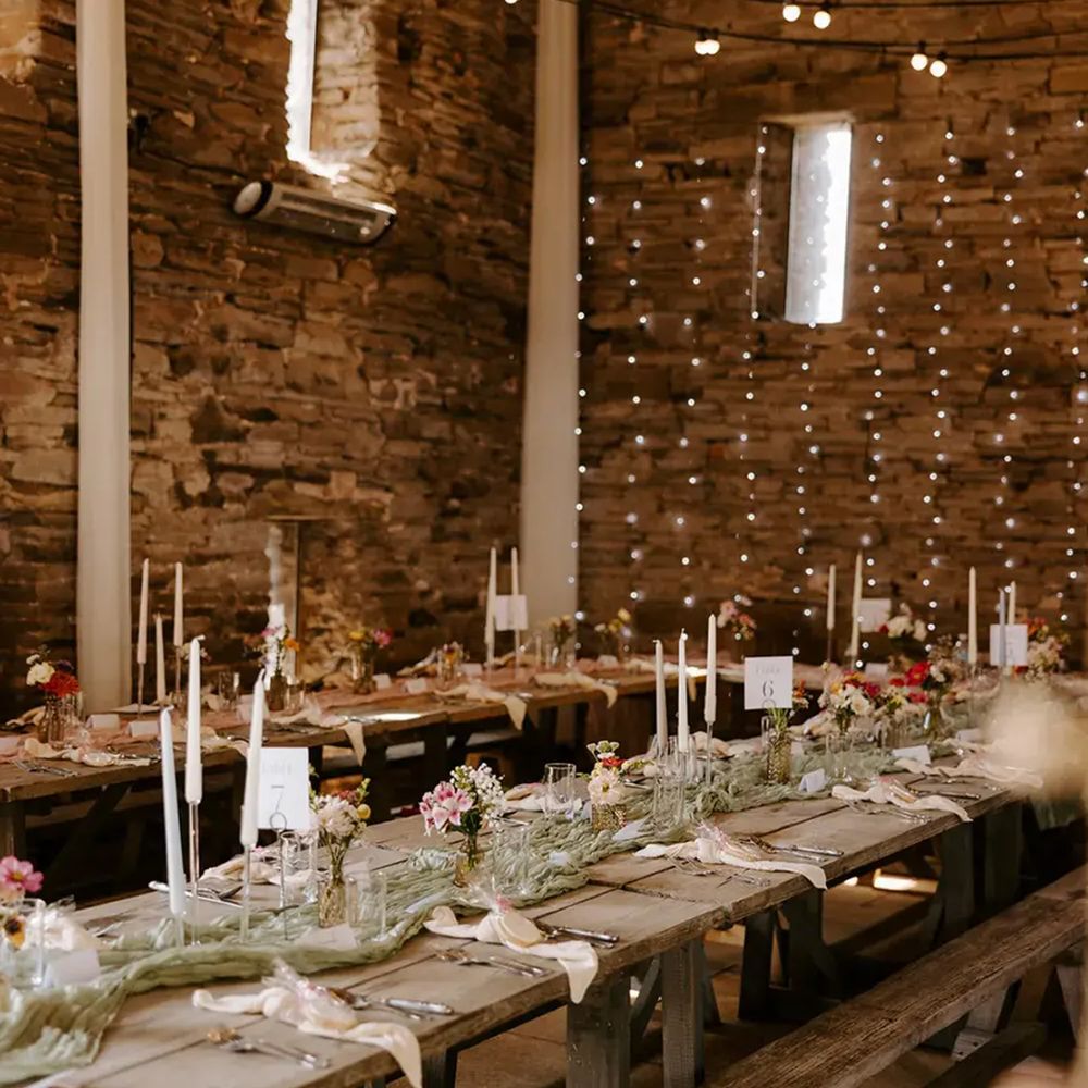 Rustic barn reception with wooden trestle tables, taper candles and fairy lights on stone walls