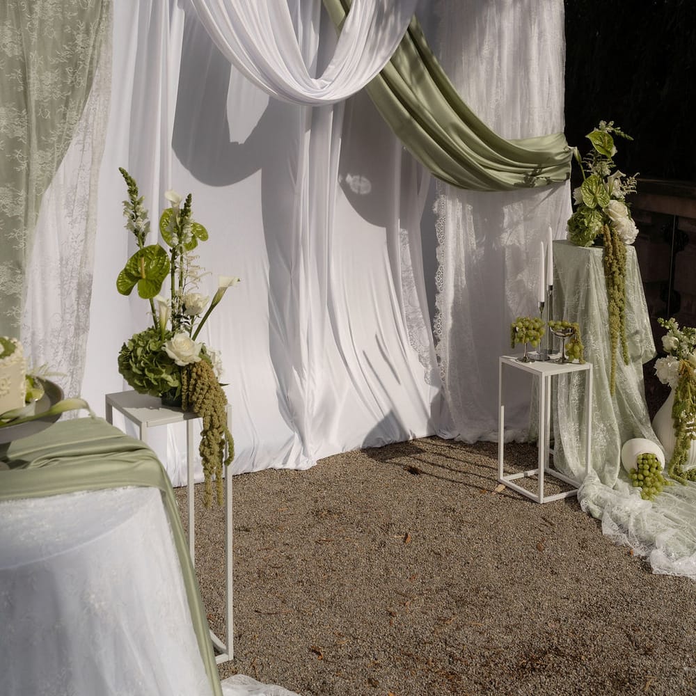 Outdoor ceremony backdrop with white and sage green draping and white floral arrangements.