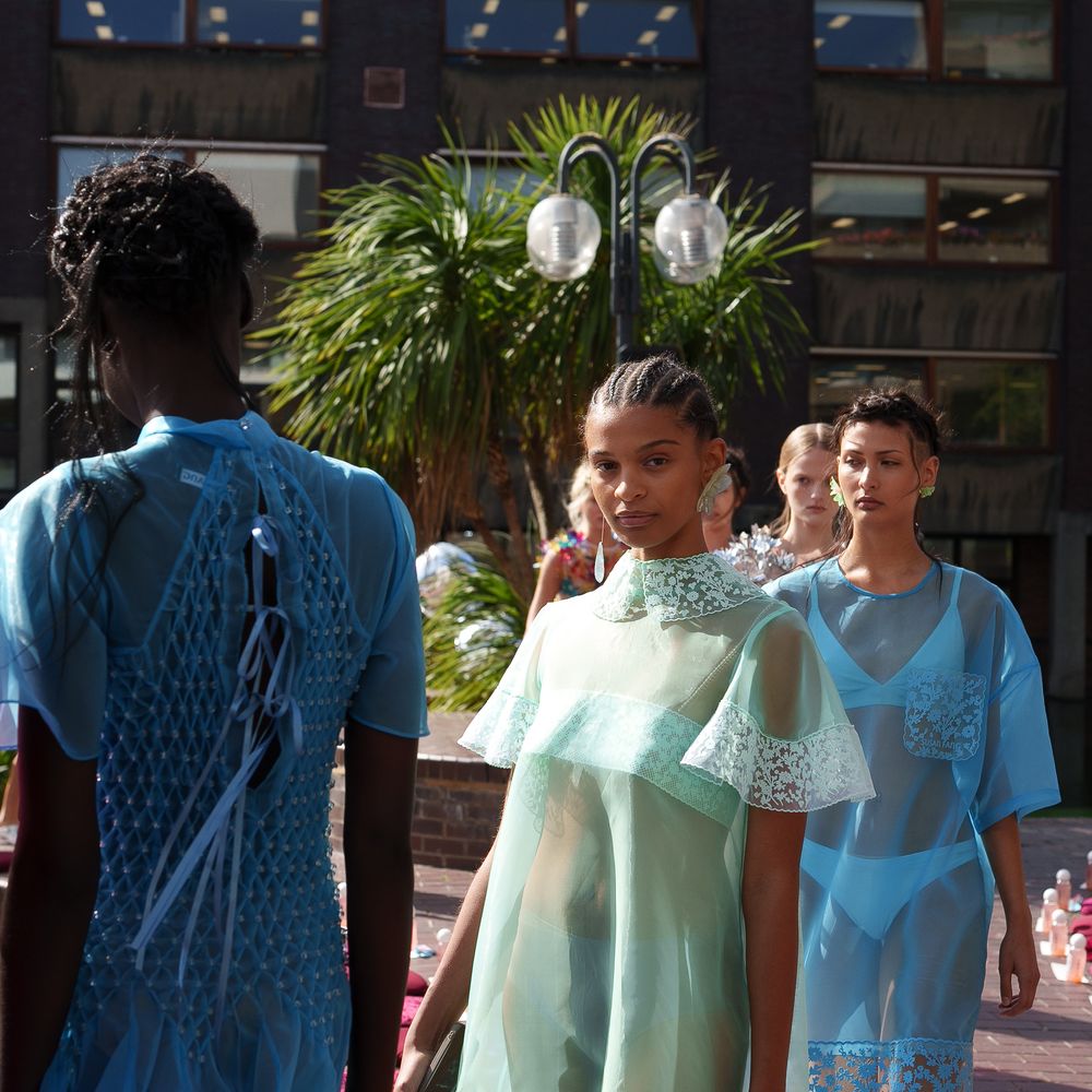 Models in sheer mint and blue lace-trimmed dresses walk an outdoor runway lined with palms.