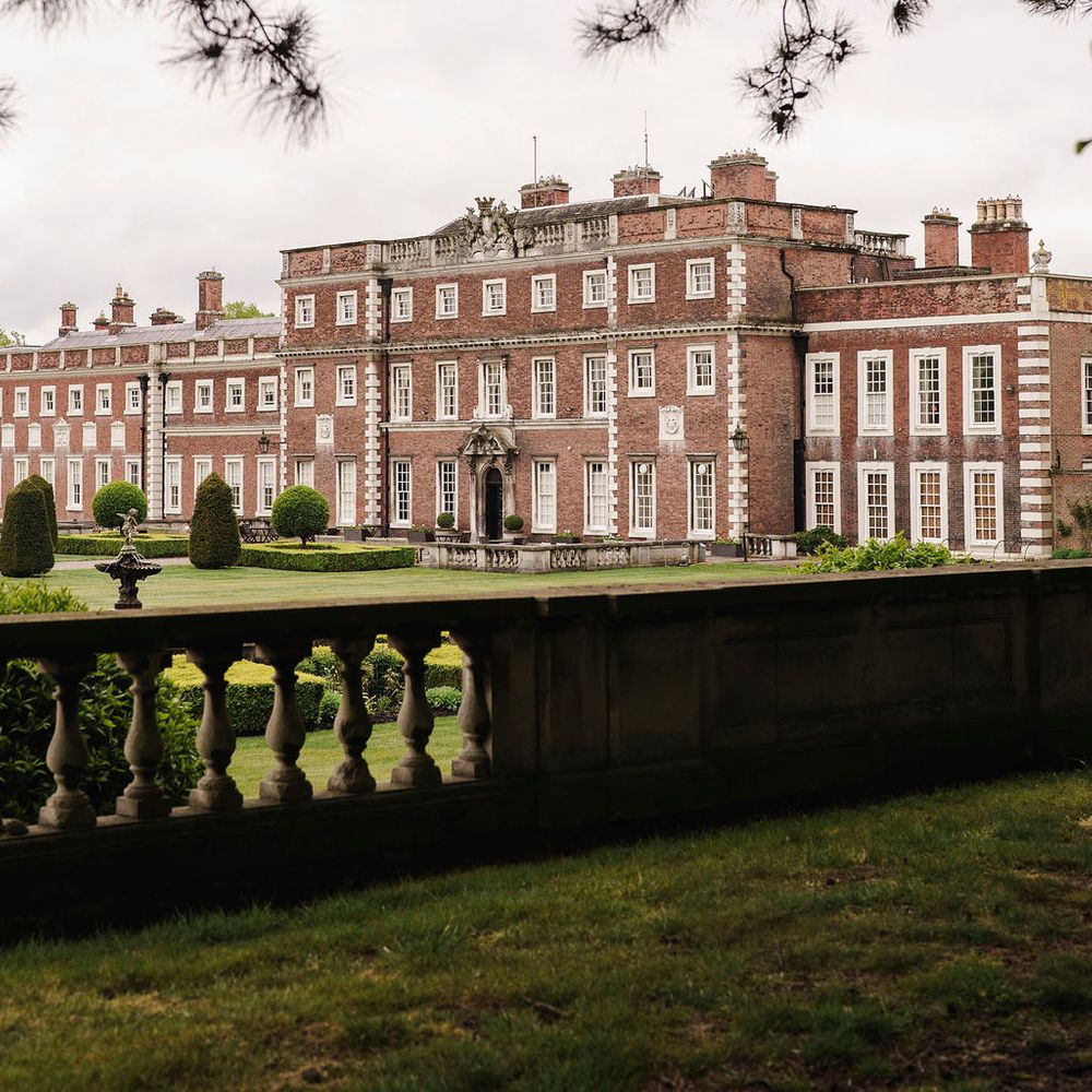 Grand red-brick stately home with formal gardens and stone balustrade, framed by trees.