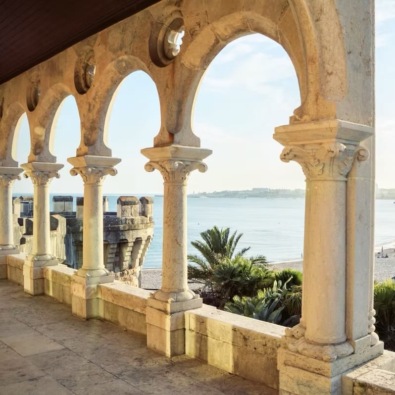 Stone archways and columns framing a coastal view of the sea, beach and palm trees at sunset
