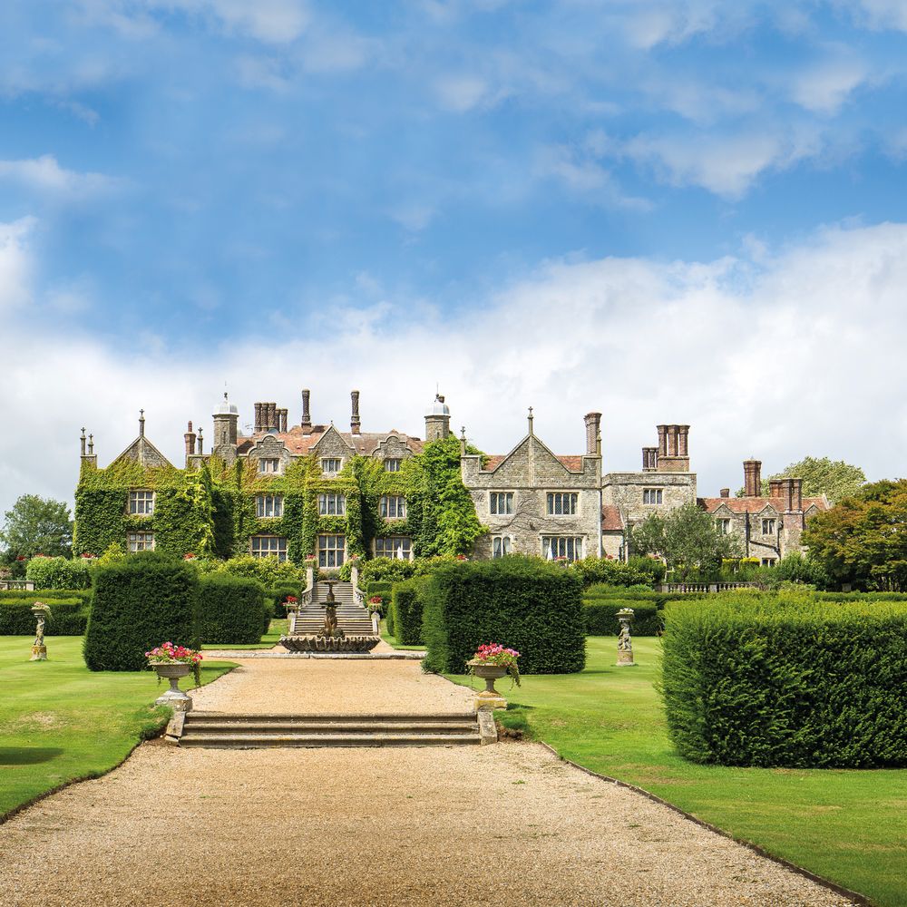 Ivy-clad stone manor house with formal gardens, clipped hedges and gravel path under a blue sky