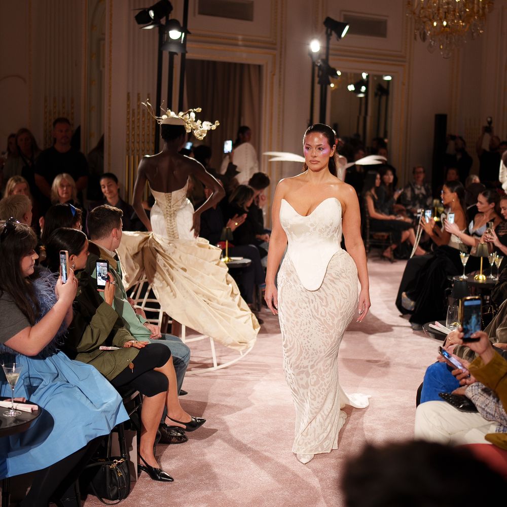 Model walking a runway in a strapless corseted ivory gown before a seated audience filming on phones