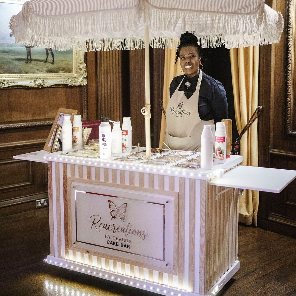 Smiling vendor behind a striped fringed cake bar cart with fairy lights in a panelled room