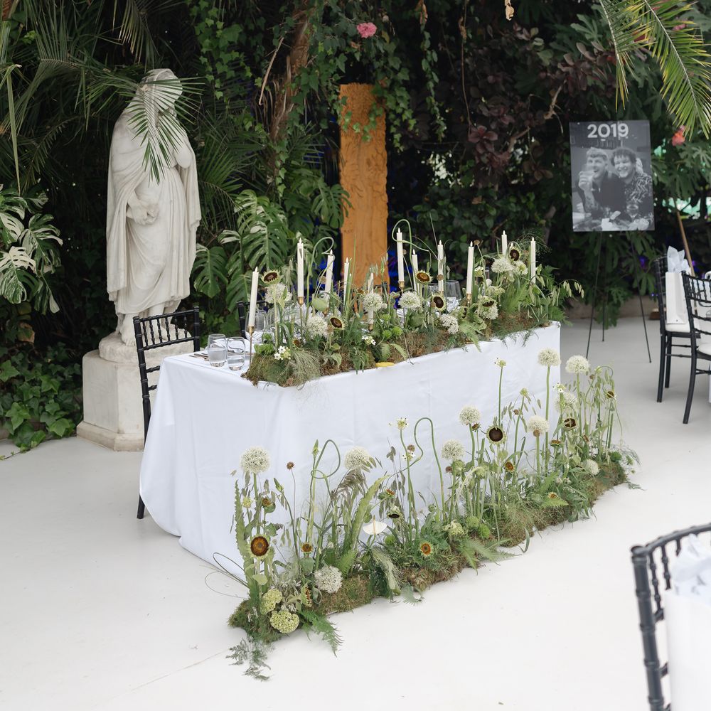 Sweetheart table draped in white linen with wildflower florals beneath tropical palms and a marble statue